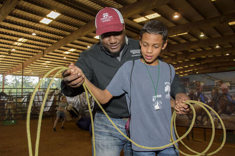 A male African American Extension agent teaches a young African American male FARMtastic visitor how to properly hold a lasso.