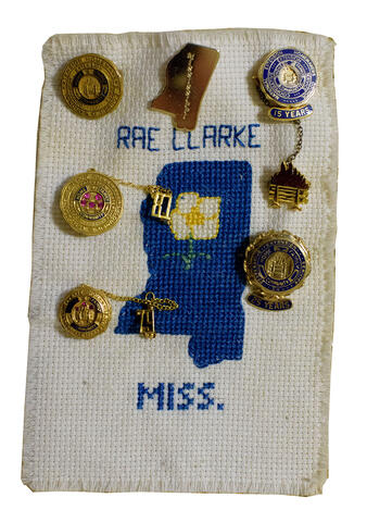 A rectangular piece of white fabric has a blue outline of the state of Mississippi and the words &quot;Rae Clarke&quot; and &quot;Miss.&quot; The fabric is decorated with several gold pins.