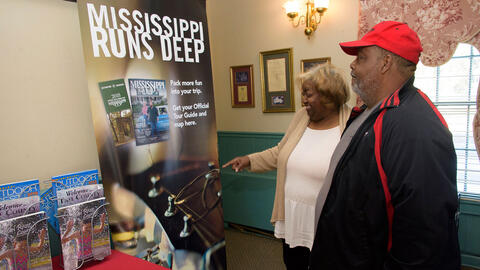 A man wearing a red hat stands next to a smiling woman pointing at a poster that says "Mississippi Runs Deep."