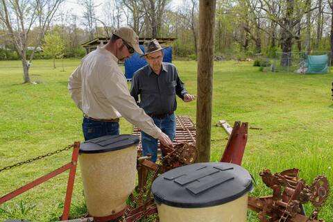 A man wearing a baseball hat and long-sleeved polo points to a metal gear on a piece of farm equipment. A man wearing a camouflage wide-brimmed hat examines the gear.