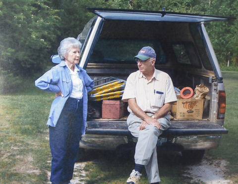 An old photo of a woman standing next to a truck with a man sitting on the bed of the truck with his legs crossed.