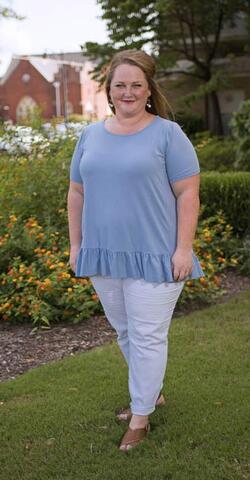A woman wearing a light blue shirt and white pants stands in green grass in front of flowery bushes.