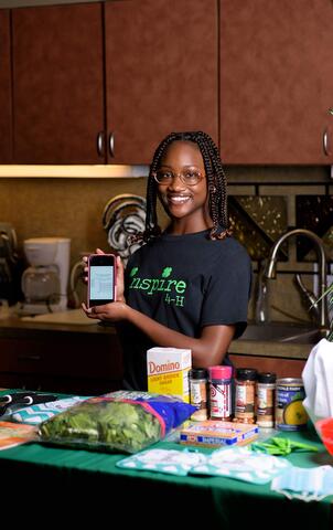 A smiling teenage girl stands behind a table covered in ingredients for a recipe displayed on a phone screen she holds in front of her.