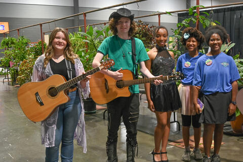 Two students, each holding a guitar, stand near a student in a party dress and two other students both wearing blue shirts.