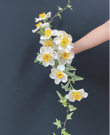 Hand holds a long S-shaped bouquet of yellow and white flowers with green leaves.