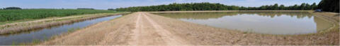 Landscape with crops growing on the left, a TWR ditch filled with water, a dirt road, and a pond-like area on the right.