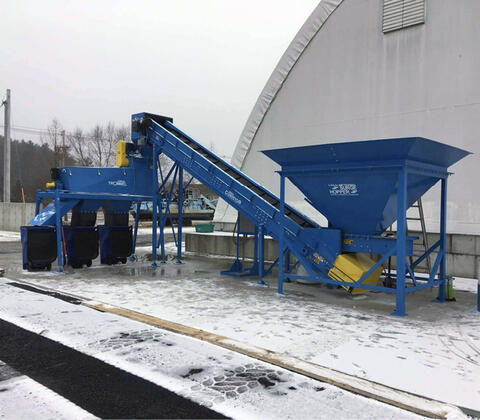 Large machinery featuring a funnel-like opening and conveyor connected diagonally to the system next to an arched building.