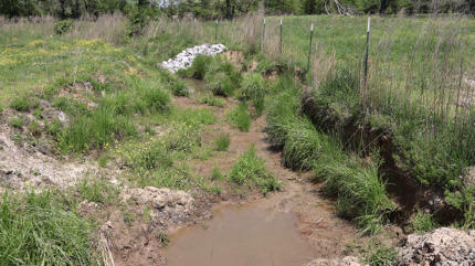 Figure3B after: A ditch features shrubs and minimal water from a drainage pipe. A fence stands on the right of the ditch. Farther down the ditch, a wide strip of rocks crosses it.
