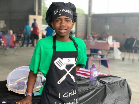 A young boy wearing a green shirt, black apron, and black chef’s hat that says “Raymond.&quot;