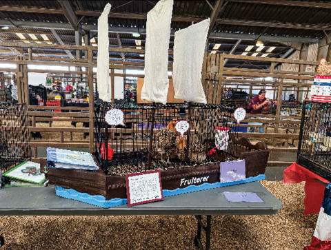 The coop is designed like a sailing ship with three white sails atop and a wooden base. Several signs adon the ship.