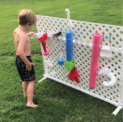 A young boy pours a bottle of water into a funnel attached to a white lattice with pool noodles, pipes, and baskets affixed to it.