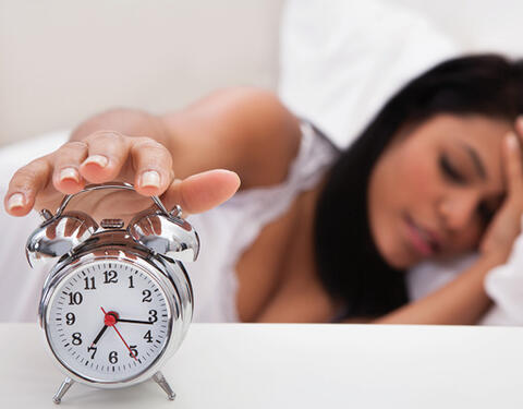 A person lying in bed reaches for the snooze button on an alarm clock.