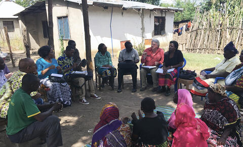 A group of people sit in a circle outside, some in chairs and some on the dirt ground. Several people are taking notes, and one person is speaking.