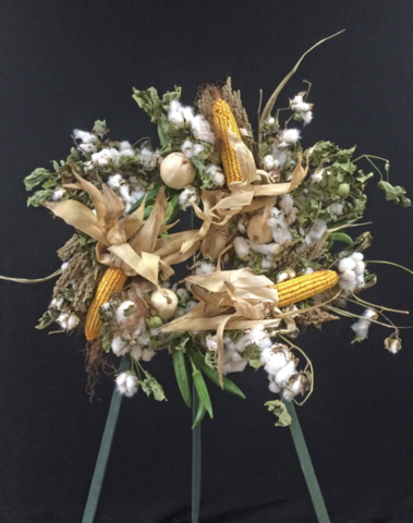 A finished wreath on a tripod display. The wreath made of stems of cotton; ears of corn and their husks; bunches of okra pods; small, white pumpkins; and stems of milo.