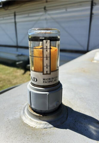 A gauge features a glass tube containing a level of liquid and is attached to a generator fuel tank outside a poultry house.