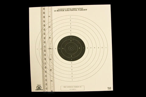 National Rifle Association 10 meter Air Pistol Target with six white outer circles and five smaller inner black circles with a ruler lying vertically on the left of the target.