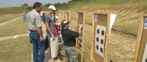Two men watching one other man set up a target sheet with six targets on a board with a number labeled 49 in the top left corner and an identical board and target is to the right of them but labeled with 50 in the top left corner.