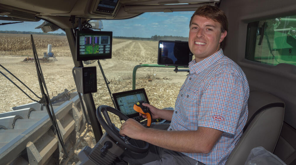 A smiling person seated in a combine in a corn field.