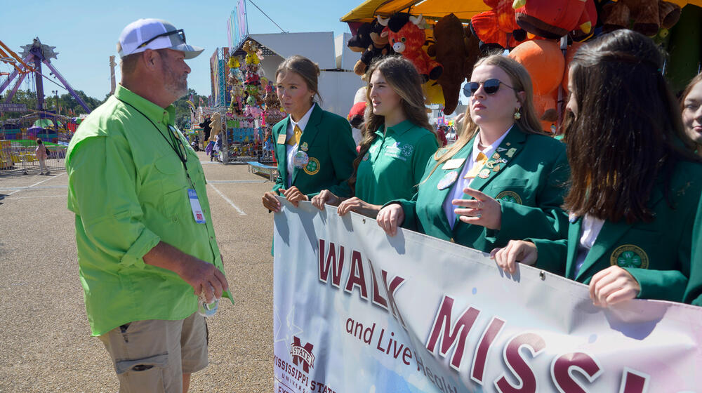 A person wearing a green polo talking to young people wearing green and holding a Walk Mississippi banner