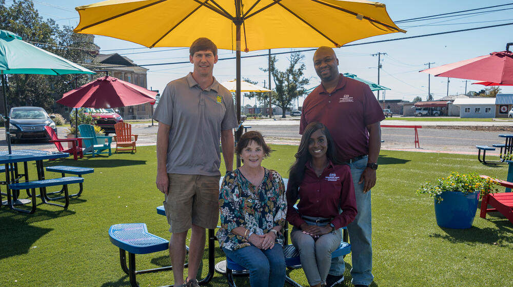 Two people standing and two people sitting under a yellow parasol.