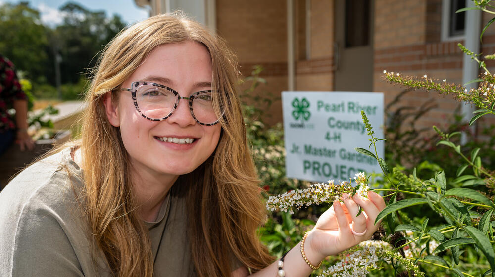 A person smiling, holding a plant in front of a sign listing, “Pearl River County 4-H Jr. Master Gardener Project.”