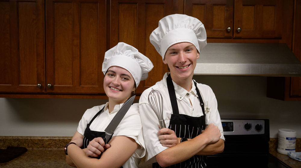 Two young people wearing chef's hats, with one holding a knife and one holding a whisk, standing back to back and smiling.