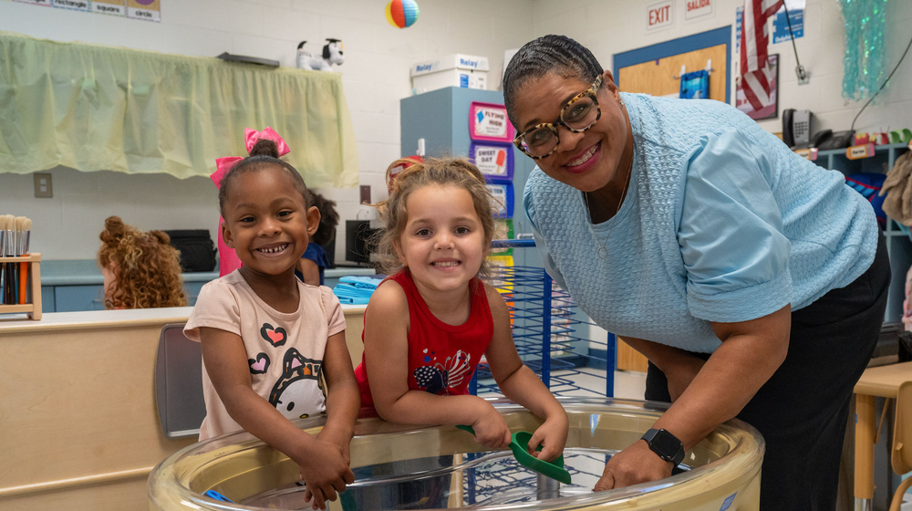 A smiling adult with two smiling children at a bead and building station.