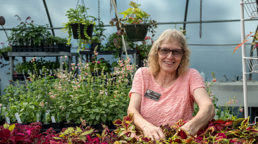 A person smiling in a greenhouse surrounded by colorful foliage.