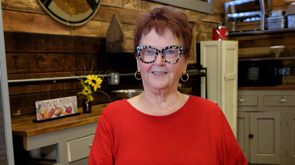 An elderly woman wearing animal print glasses and a bright red shirt stands smiling. Photo credit: Kevin Hudson