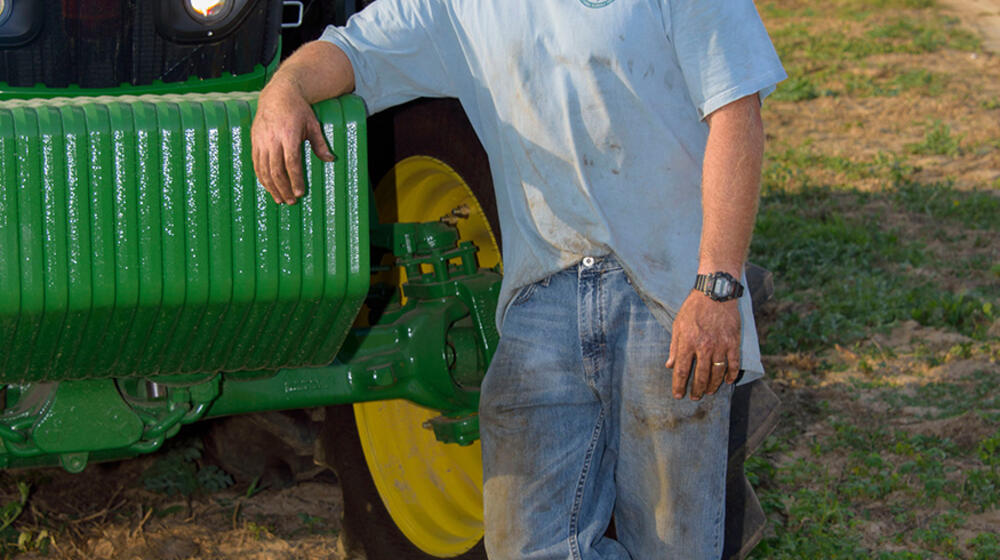 A man wearing a blue t-shirt and blue jeans leans against a green tractor.