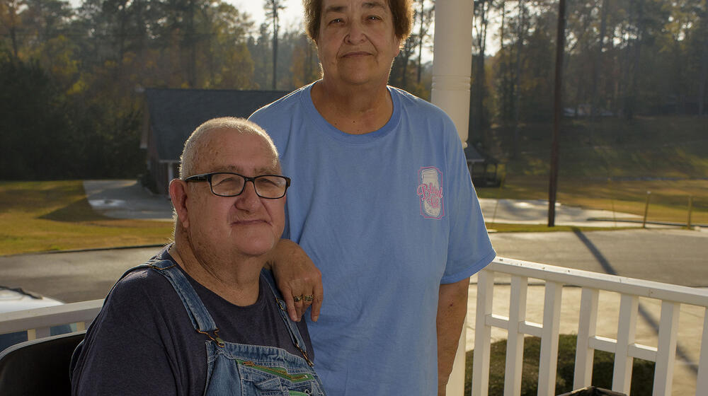 A gray-haired man wearing overalls and glasses sits beside a raised salad table, and a black-haired woman stands behind him, wearing a blue T-shirt, with her hand on his shoulder.