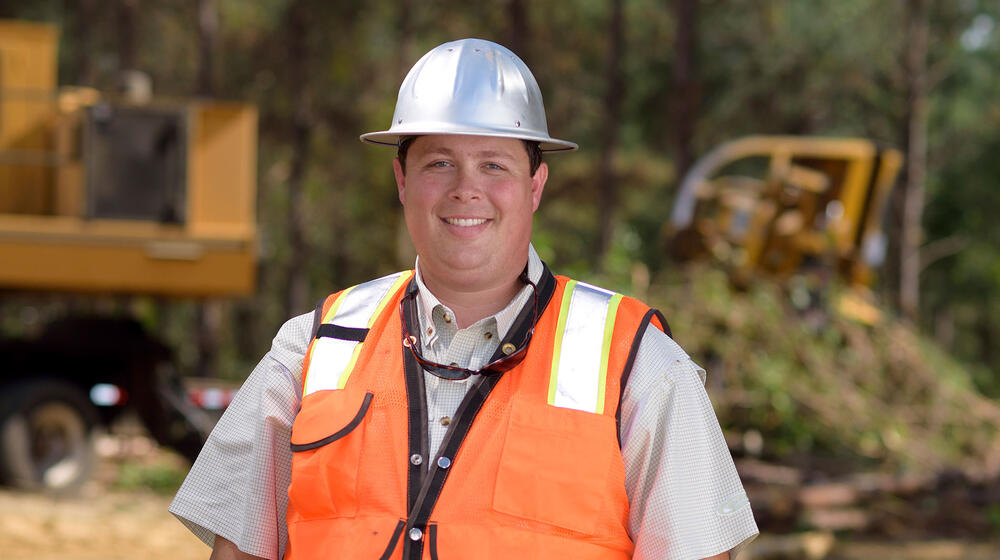 A young man with a metal construction hat and bright orange vest stands in front of his work site with his hands tucked into his blue jeans.