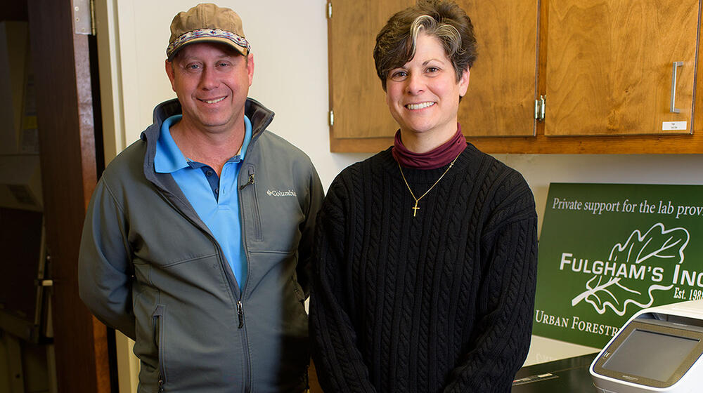 A man wearing a light blue collared shirt and a grey zip-up jacket stands next to a short-haired woman wearing a black sweater.