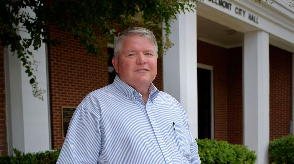 A man wearing a light blue and white striped shirt and blue jeans stands in front of Belmont City Hall.