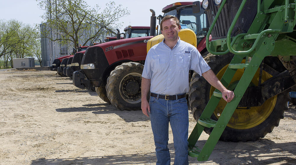 A man wearing a blue collared shirt rests his hand on a green tractor with several red tractors parked behind him.