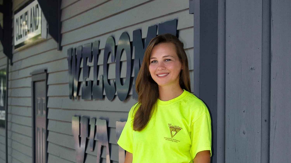 A teen with brown hair and wearing a green Junior Master Wellness Volunteer T-shirt stands in front of the Cleveland, Mississippi, Welcome Center.