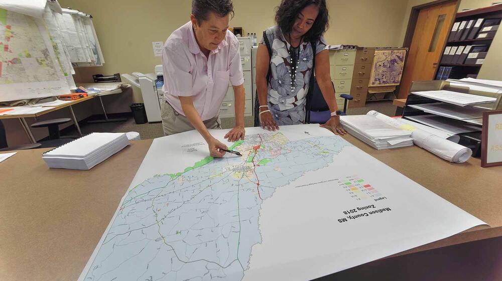 On the left, a woman with short hair and a light pink, collared shirt leans on the table, holding a pen above an area of a map. On the right another woman wearing a blue floral dress leans on the table and looks at it.