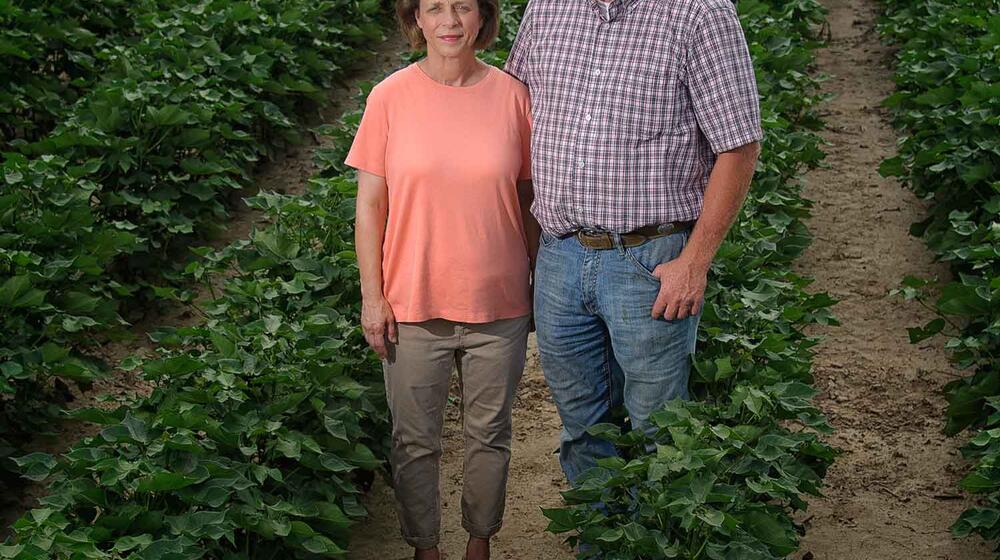 A couple stands among crop rows. The woman, left, has short brown hair, a pink shirt, and khaki pants, and  a taller man wears a white and tan baseball cap, a checkerboard-striped shirt, and blue jeans.