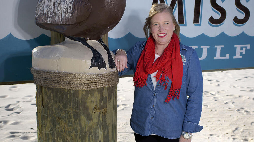 A smiling woman with blonde hair, a red scarf, a denim shirt, and black pants rests her arm on the side of a “Welcome to the Mississippi Gulf” sign.
