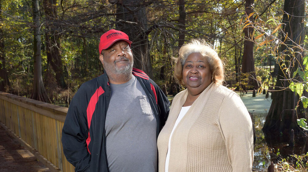 A couple, a man with a black and red jacket and a woman with a tan cardigan, stands next to a wooden railing located outside with trees and water in the background.