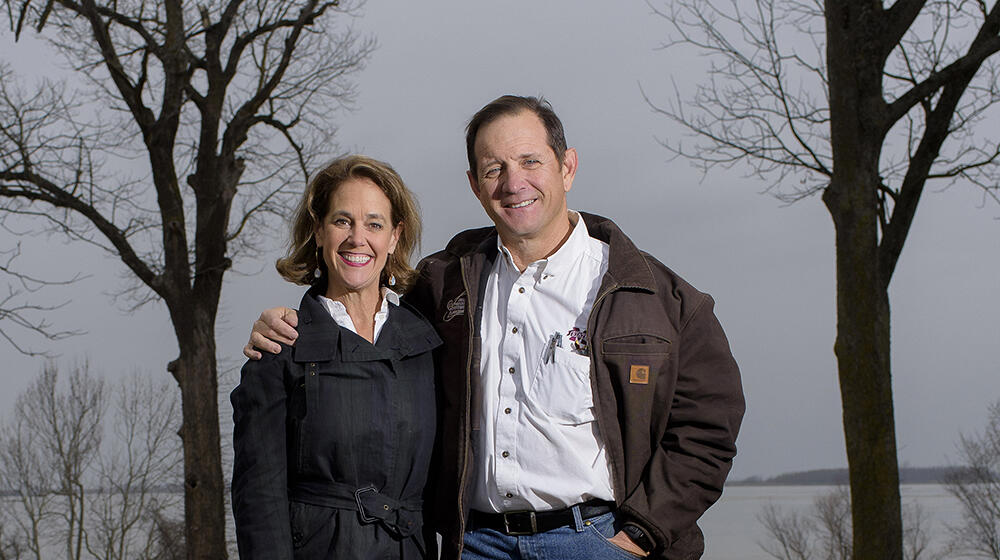 A man stands next to a woman with his arm around her shoulders while they both smile at the camera; a lake rests in the background.