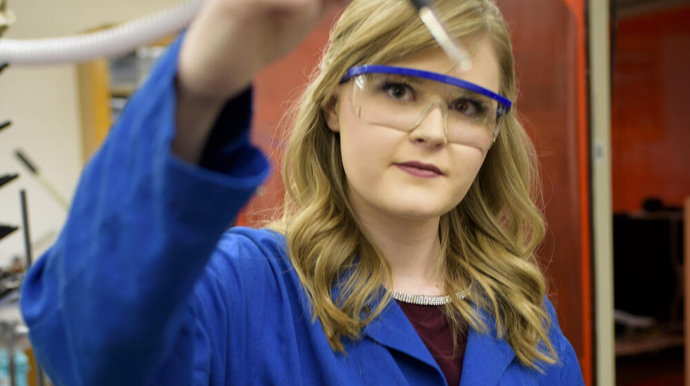 A blonde young woman wearing a blue lab coat and safety glasses holds a glass dropper up to the camera.