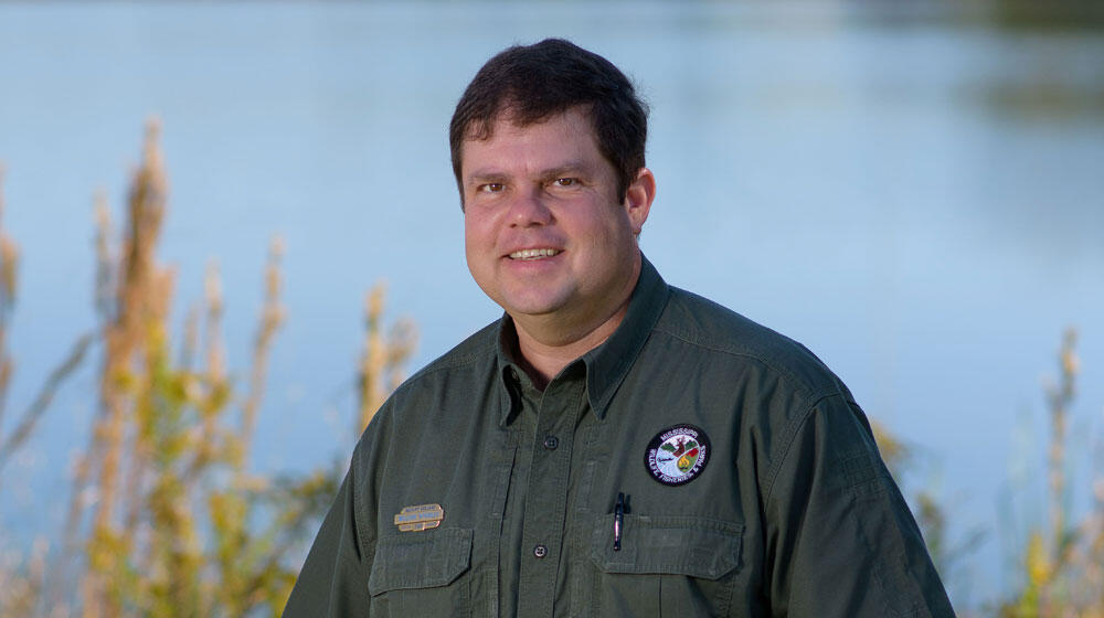 A man wearing a green collared shirt stands smiling.