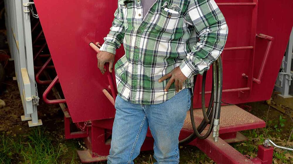 A man wearing a green and white plaid shirt, blue jeans, and a brown baseball cap stands in front of a bright red machine parked inside a metal building.  