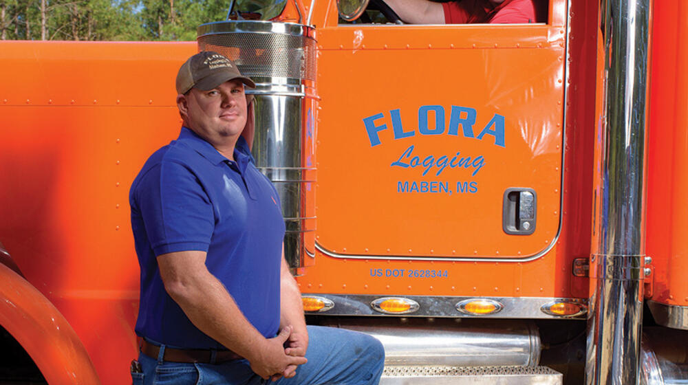 A man in a blue shirt stands in front of an orange semi-truck with another man in the driver seat.