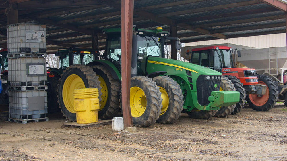A green tractor, flanked on the right by a smaller red one, stands beneath a metal roof.