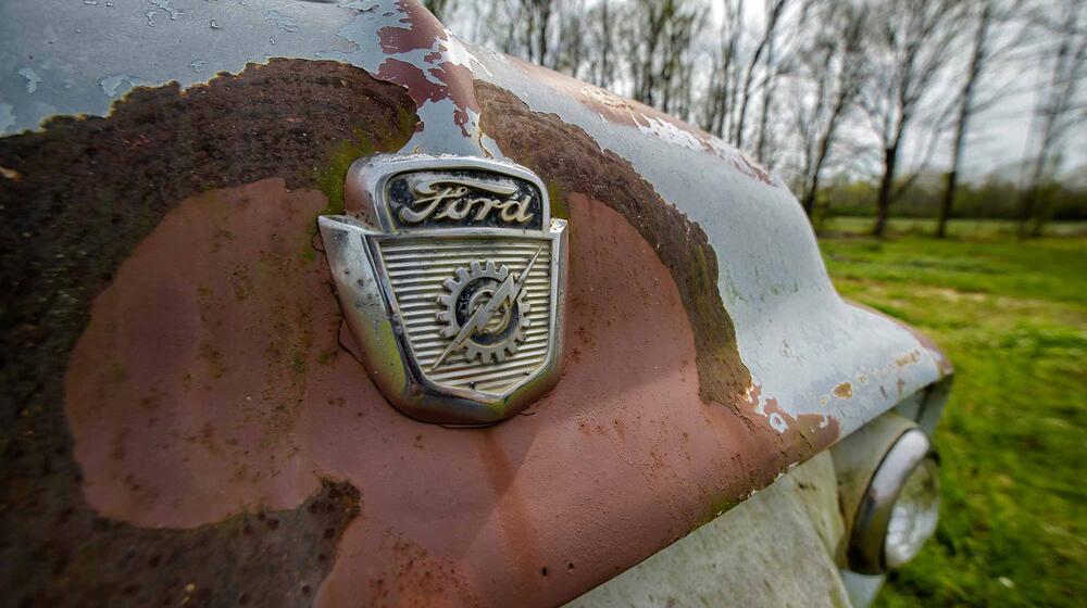 The front of an old Ford truck with the logo centered in an orange-colored rusty area of the light blue truck. 