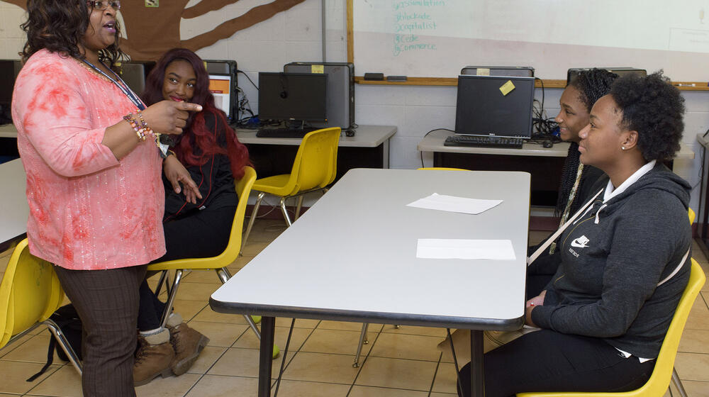 A woman, leftmost, wearing a coral blouse and brown slacks, points and talks to two teens sitting in yellow chairs at a gray table.