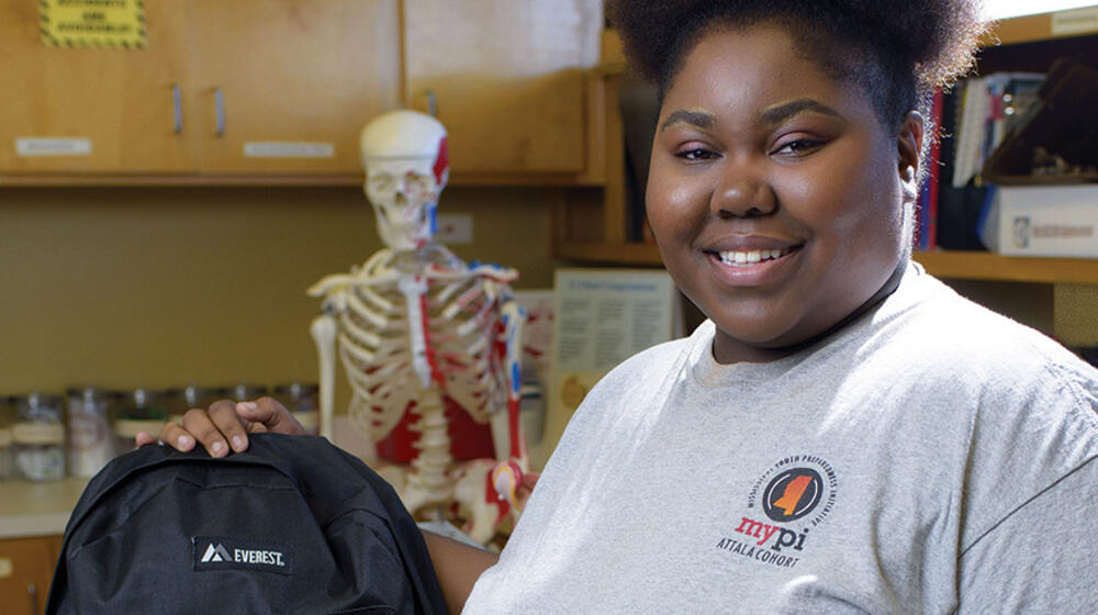 A smiling teenage girl standing next to a black backpack.