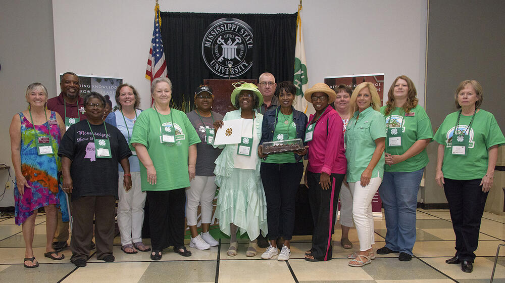 A group of 14 men and women stand on either side of a woman wearing a bright green hat and holding up an award.